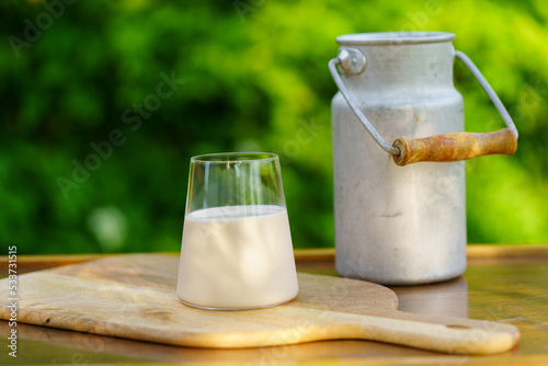 A glass of milk and an old metal milk can on a wooden table against the green defocused natural background.