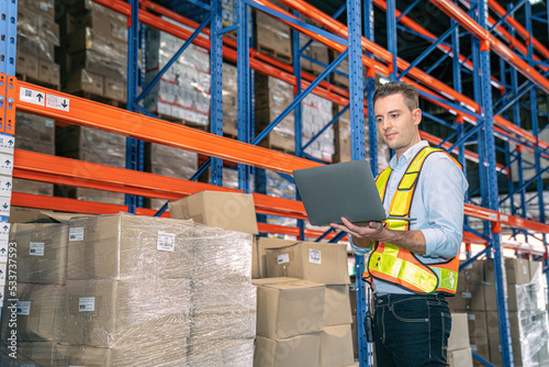Smart smiling caucasian warehouse man,worker,manager use laptop to check goods on shelf for product distribution stock management, Logistics shipping business planning concept with copy space