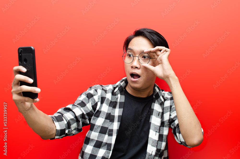 Asian man looks surprised at screen of mobile phone. happy asian man with gesture while holding phone. wow expression. asian man with glasses holding phone. Asian man looks surprised at screen of mobile phone. happy asian man with gesture while holding phone. wow expression. asian man with glasses holding phone.