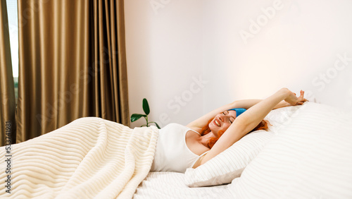 Top view portrait of smiling Caucasian young woman in sleeping mask relaxing on cozy bed at home or hotel, happy millennial girl look at camera rest in comfortable bedroom, relaxation concept