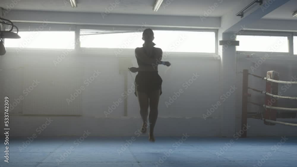 Sportswoman skipping rope, intensive cardio training in gym, front view wide shot. Young professional fighter silhouette exercising, jumping Sportswoman skipping rope, intensive cardio training in gym, front view wide shot. Young professional fighter silhouette exercising, jumping