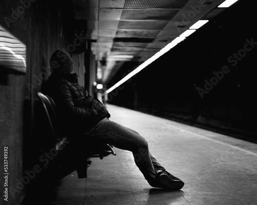Grayscale side view of man sitting on bench in the platform waiting for train