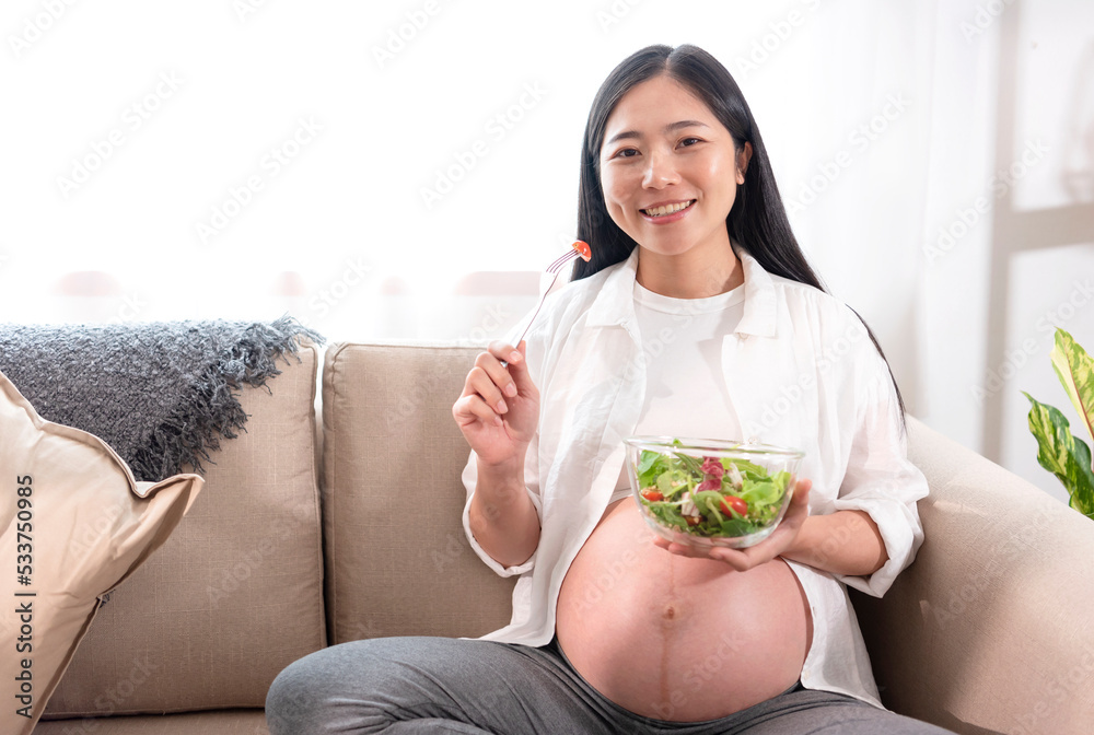 Happy asian pregnant woman eating salad on sofa at home. eating many different vegetables during pregnancy, healthy pregnancy concept. Happy asian pregnant woman eating salad on sofa at home. eating many different vegetables during pregnancy, healthy pregnancy concept.