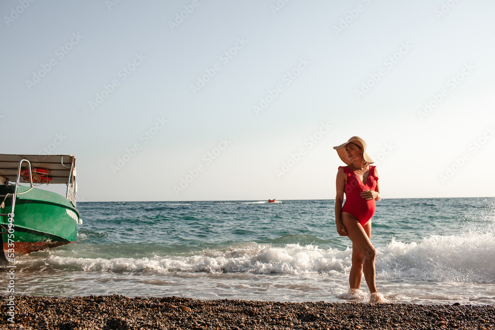 Beautiful slender pregnant woman is resting on the beach, the girl is dressed in a red solid swimsuit and straw hat, sitting under umbrella