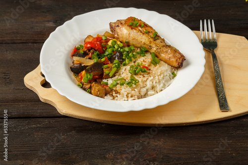Rice with vegetables and chicken leg in a white plate on a handmade wooden board next to a fork on a wooden table.