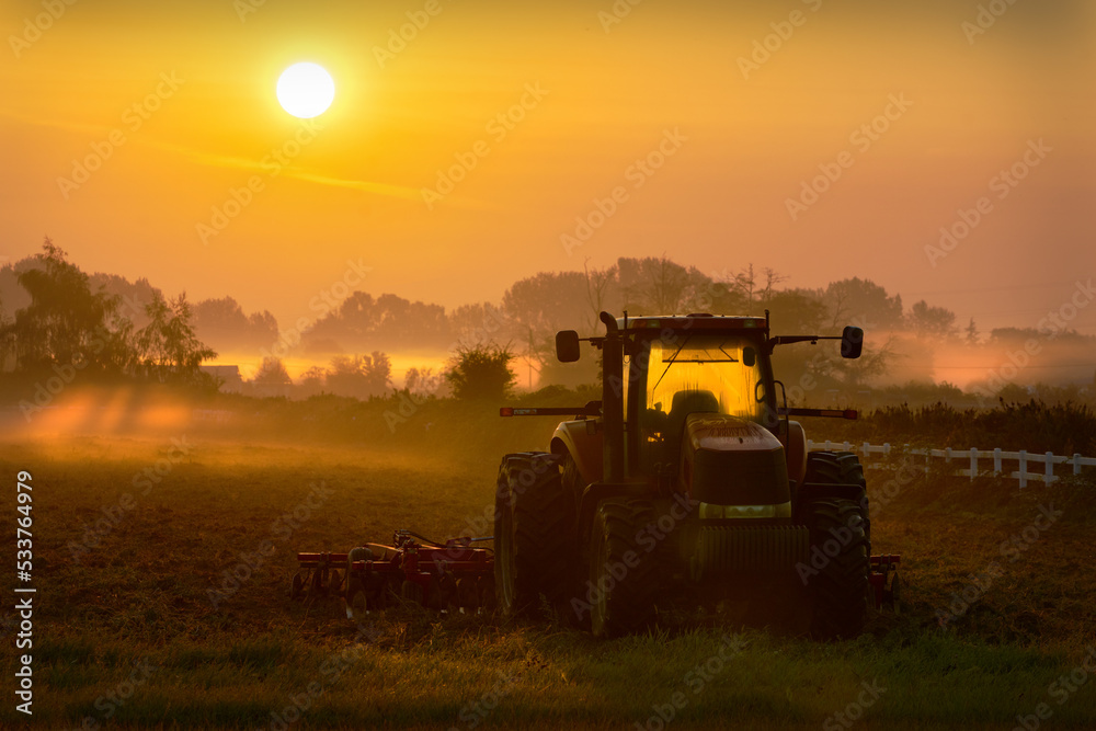 Fototapeta premium Foggy Morning Tractor Sunrise. Sunrise over a misty farm field and tractor.