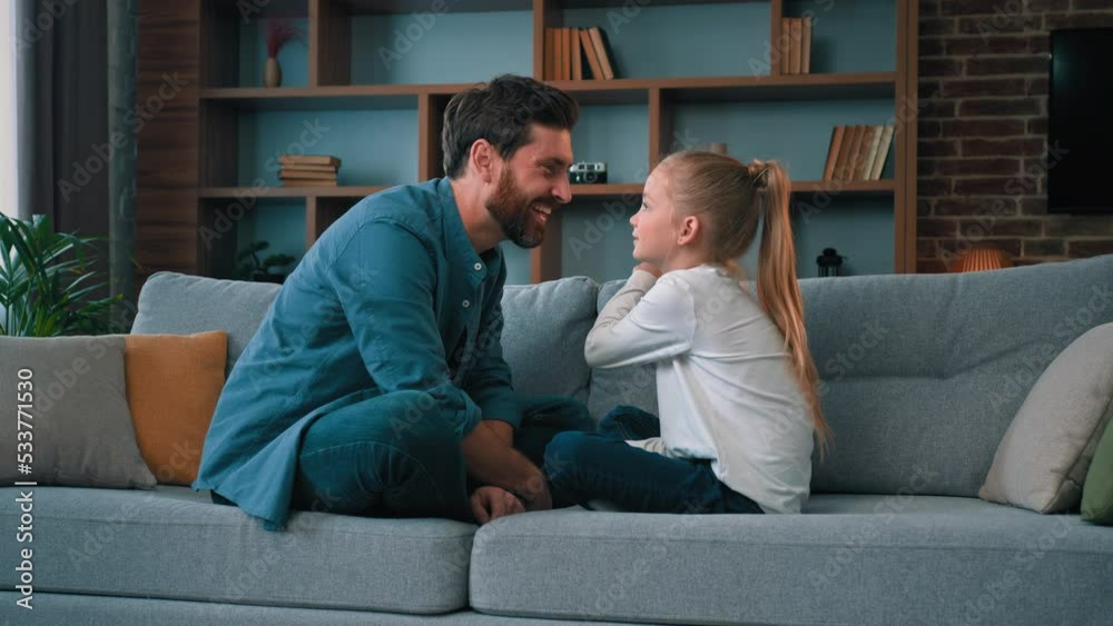 Playful caucasian young family daddy and small daughter sitting comfortable on couch loving cute girl touching father nose funny joke playing game with parent having fun spending free time together Playful caucasian young family daddy and small daughter sitting comfortable on couch loving cute girl touching father nose funny joke playing game with parent having fun spending free time together