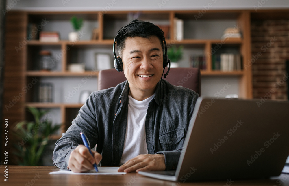 Cheerful middle aged chinese man in headphones has video call on pc, makes notes in cabinet interior Cheerful middle aged chinese man in headphones has video call on pc, makes notes in cabinet interior