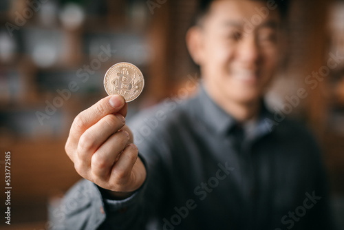Smiling successful adult chinese man show bitcoin coin, manager work with cryptocurrency in office interior