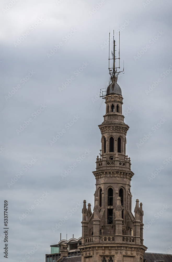 Antwerpen, Flanders, Belgium - July 10, 2022: Along Scheldt River. Beige-brown stone Historic Loodswezen, shipping pilots, steeple part with antennas on top under gray cloudscape