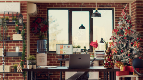 Festive decorated workplace with laptop on desk, christmas tree and lights as winter season decorations. Empty office filled with seasonal ornaments used to celebrate xmas holiday.