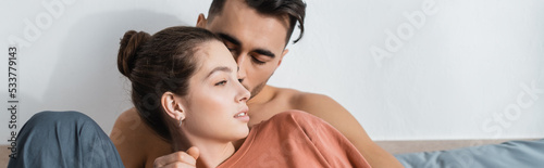 shirtless man kissing girlfriend in t-shirt looking away in bedroom, banner.
