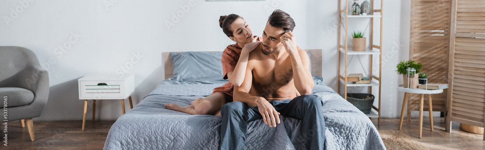 sexy man in pajama pants near girlfriend embracing him on bed at home, banner. sexy man in pajama pants near girlfriend embracing him on bed at home, banner.
