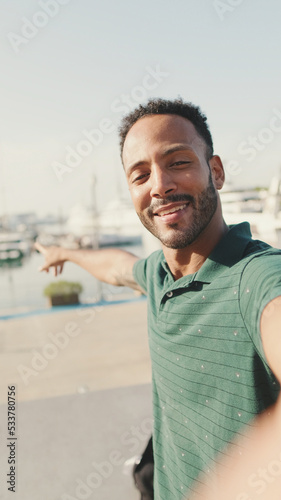 Happy young man making video call from phone while sitting in harbor with yachts on background