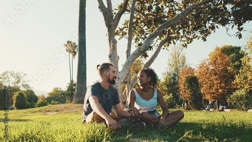 Happy smiling interracial couple talking while sitting on the grass in the park. Backlight