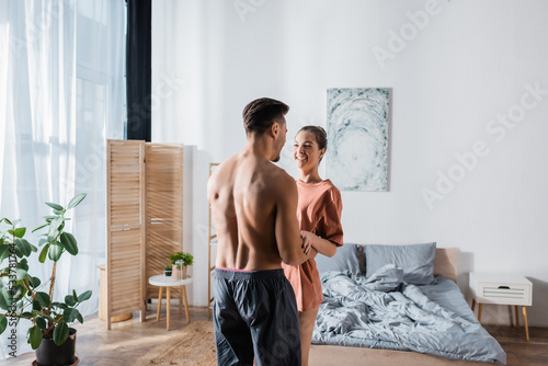 cheerful woman in t-shirt laughing near shirtless boyfriend embracing her in bedroom.