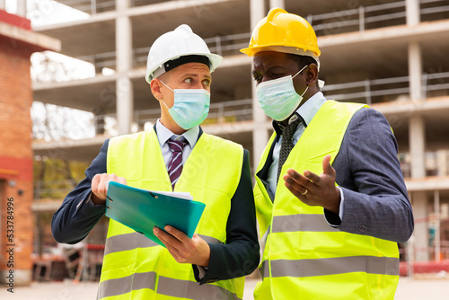 Two engineers in protective masks, working on a construction site during a pandemic, discuss a construction plan, holding an ..estimate in their hands