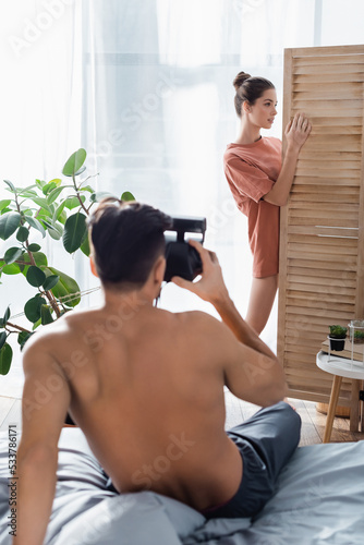 seductive woman posing near room divider while shirtless man with vintage camera taking photo on blurred foreground.