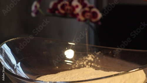 Man putting salt over mix of yeast and flour in glass bowl with warm yellow light and orchid in background photo