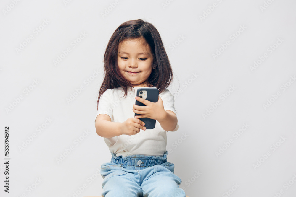 a cute little girl of preschool age is standing on a white background in a white T-shirt, thoughtfully sitting on a chair and looking at the phone. The theme of children's happiness a cute little girl of preschool age is standing on a white background in a white T-shirt, thoughtfully sitting on a chair and looking at the phone. The theme of children's happiness