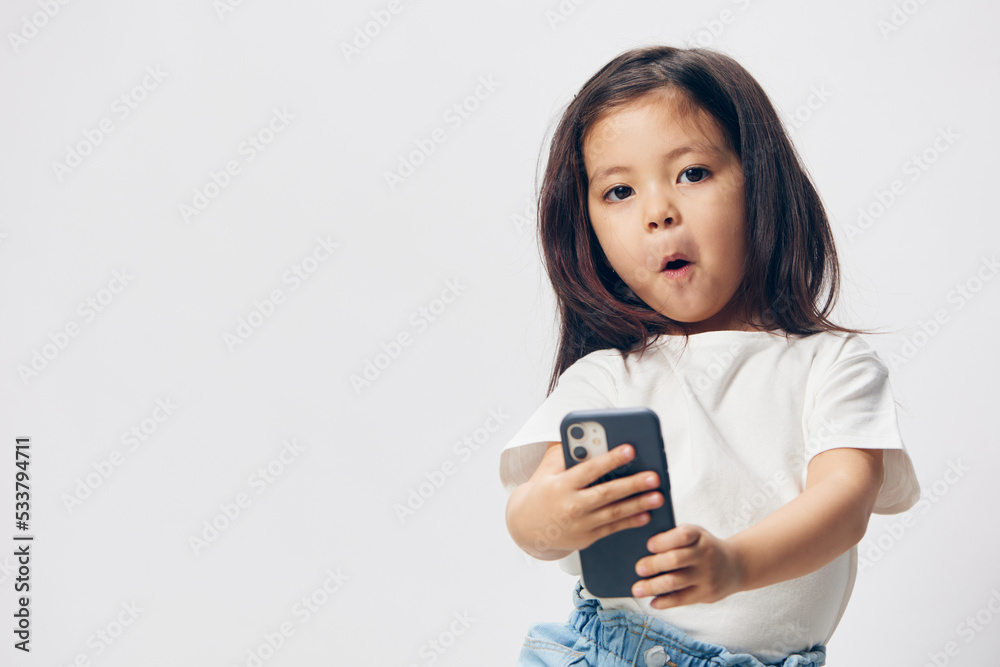a cute, happy girl of preschool age stands on a light background in a white T-shirt with a fashionable smartphone and looks into it with enthusiasm a cute, happy girl of preschool age stands on a light background in a white T-shirt with a fashionable smartphone and looks into it with enthusiasm