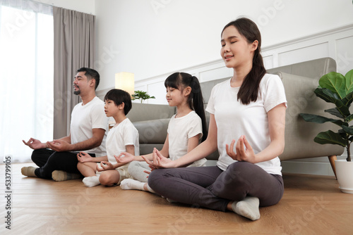 Happy Asian Thai family, parents, and children sit on living room floor, close their eyes together, tranquil meditate in lotus position, and practice healthy yoga, domestic home lifestyle weekend.