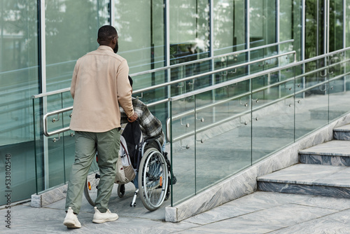 Back view of adult man assisting partner in wheelchair going up accessibility ramp in city, copy space