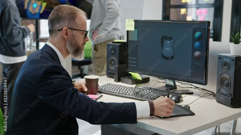 Caucasian bearded male artist designer draws on a tablet a 3D model of teapot for a computer game in the office of a project development studio. Caucasian bearded male artist designer draws on a tablet a 3D model of teapot for a computer game in the office of a project development studio.