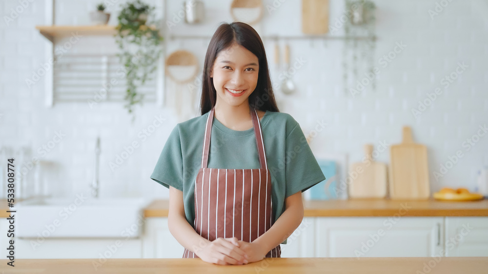 Happy young asian woman wearing apron and standing in kitchen room. Beautiful female smiling and looking at camera feeling confident Happy young asian woman wearing apron and standing in kitchen room. Beautiful female smiling and looking at camera feeling confident