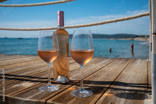 Glasses and bottle of cold rose wine from Provence served outdoor on wooden yacht pier with view on blue water and white sandy beach Plage de Pampelonne near Saint-Tropez, France