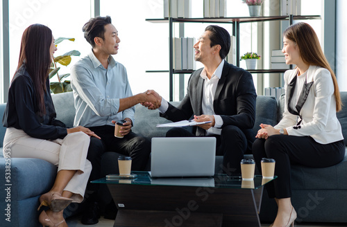 Millennial Asian professional successful businessman in formal suit sitting with female secretary shaking hands with clients after job negotitation acheivement agreement in company office living room