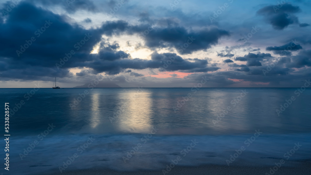 Fototapeta premium Evening twilight over the Indian Ocean. Blue clouds in the sky, highlighted in pink. Reflection on turquoise water. Foam of waves on the sand of the beach. Long exposure. Seychelles. Mahe. Beau Vallon