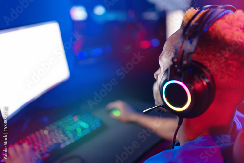 Streamer african young man rejoices in victory professional gamer playing online games computer with headphones, neon color, back view