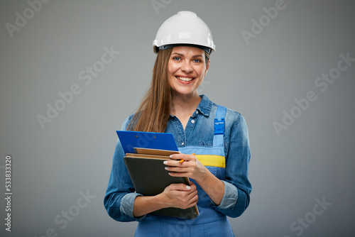 Smiling woman engineer in safety helmet and overall holding books and clipboard. Isolated female builder portrait.