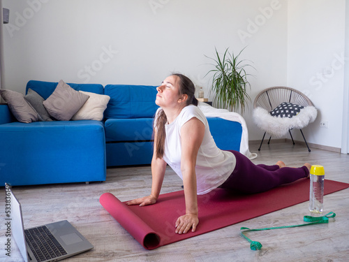 European girl looking on laptop during practising yoga. Concept of fitness, sport at home and healthy lifestyle. Online training, watching video tutorial on a laptop. Woman doing exercises on yoga mat