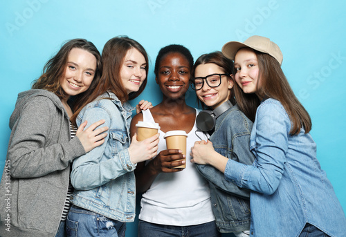 Group of young five female friends of different nationalities holding a cups of coffee to go over blue background.