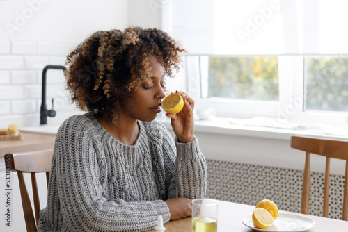 African American woman with a cold sniffs lemon to check her well-being  measures her body temperature and drinks soluble pills or vitamins  A tired sick woman at home takes pills for sore throat and