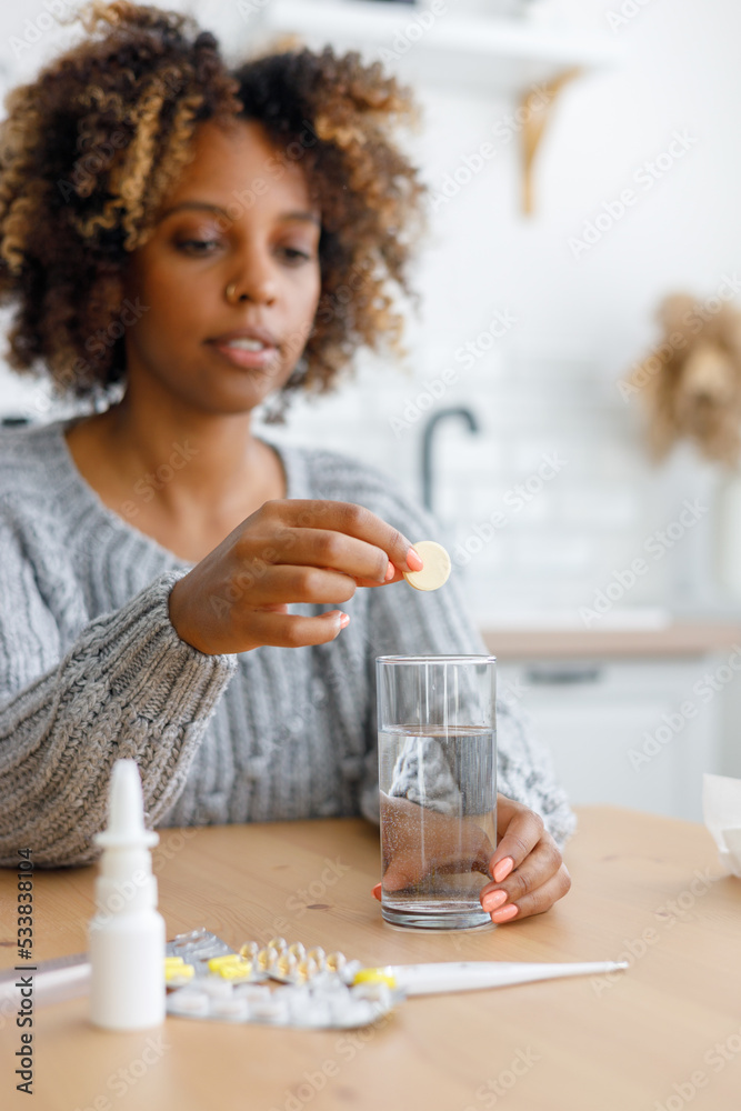 African American woman with a cold and headache drinks soluble cough pills and antipyretics. A tired sick woman at home takes pills for fever and sore throat or vitamins. African American woman with a cold and headache drinks soluble cough pills and antipyretics. A tired sick woman at home takes pills for fever and sore throat or vitamins.