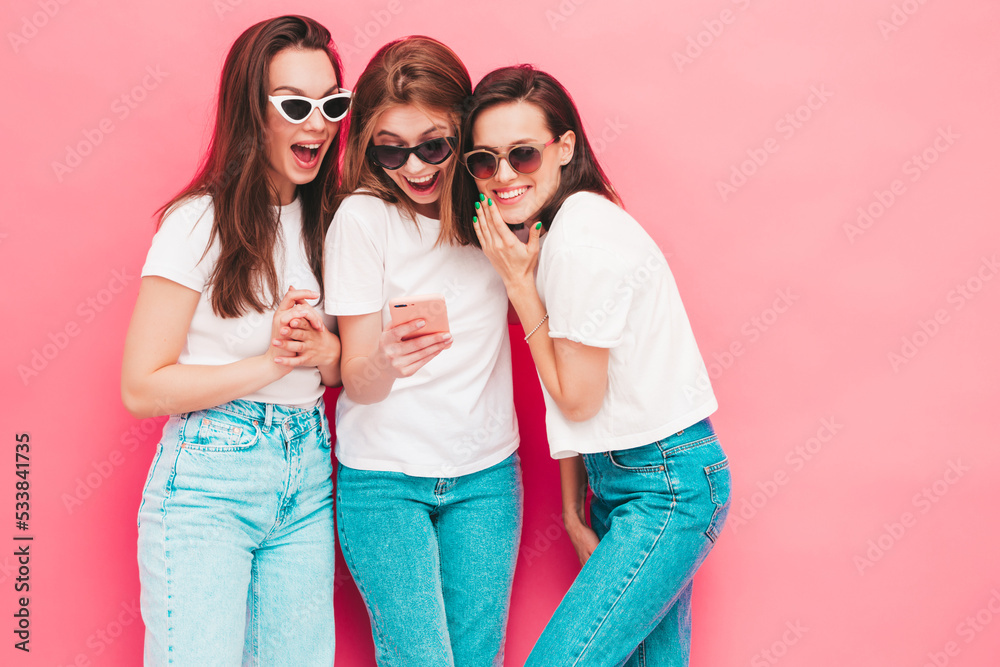 Three young beautiful smiling hipster female in trendy summer  t-shirt and jeans clothes. Sexy carefree women posing near pink wall in studio. Positive models looking at smartphone screen. Using apps