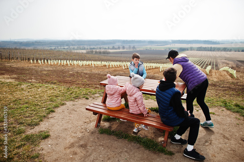 Mother with children sit at wooden table against vineyard.