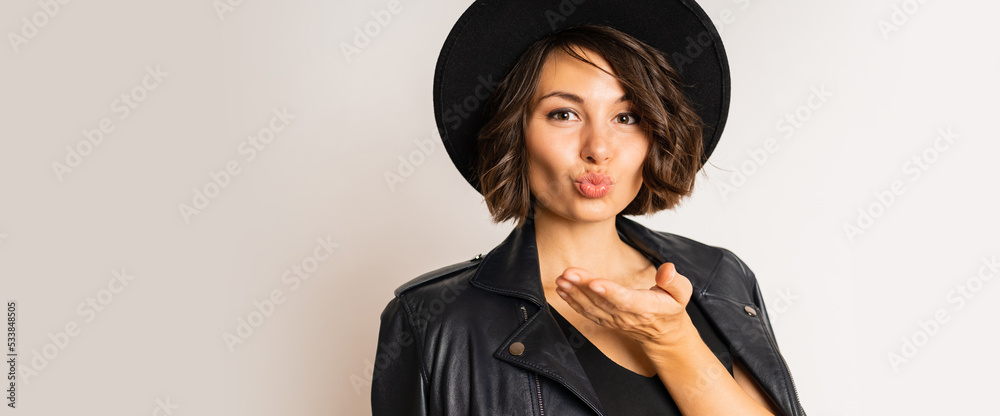 Elegant woman in black hat, evening dress and leather jacket posing on white background in studio. Elegant woman in black hat, evening dress and leather jacket posing on white background in studio.