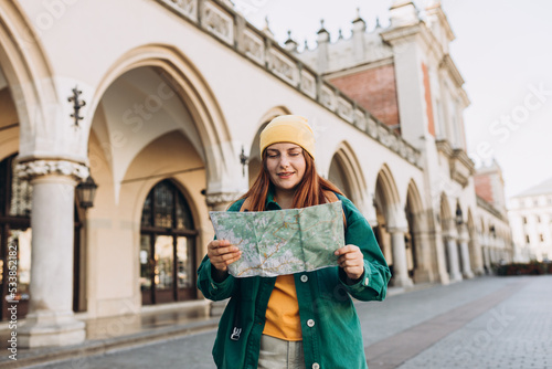 Attractive young female tourist is exploring new city. Redhead girl in hat holding a paper map on Market Square in Krakow. Traveling Europe in autumn. High quality photo