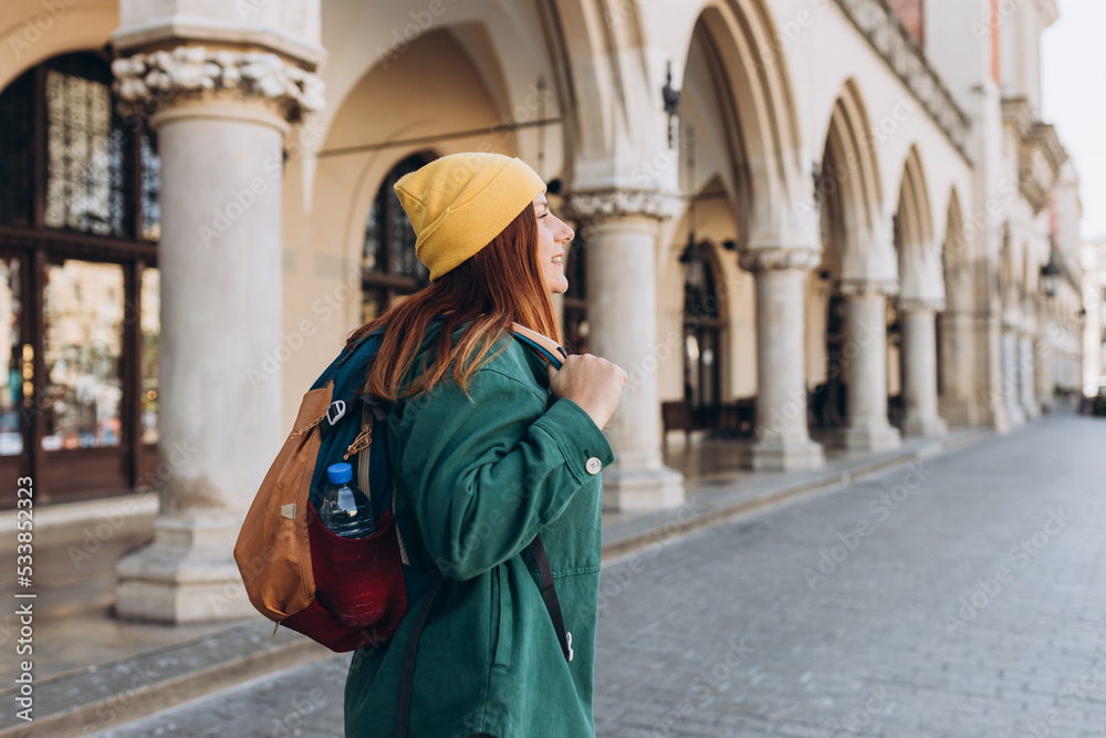 Young happy woman traveler in yellow hat with a backpack traveling Europe in autumn. Stylish female foreigner examines architectural monument during her long-awaited vacation