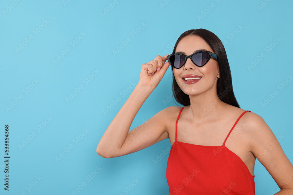 Attractive happy woman touching fashionable sunglasses against light blue background. Space for text