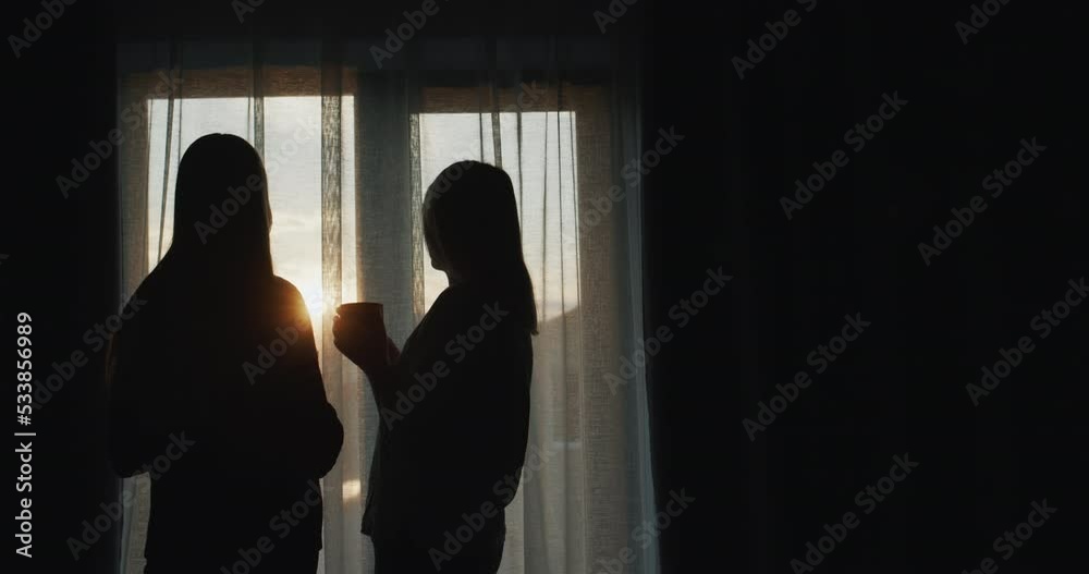 A woman with a teenage daughter stand at the window at sunset. Holding cups of tea. Rear view A woman with a teenage daughter stand at the window at sunset. Holding cups of tea. Rear view
