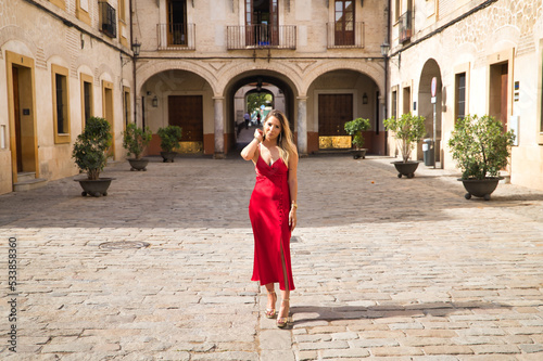 Young, attractive, blonde woman in an elegant red party dress touching her hair while strolling through the city. Concept beauty, fashion, elegance, luxury.