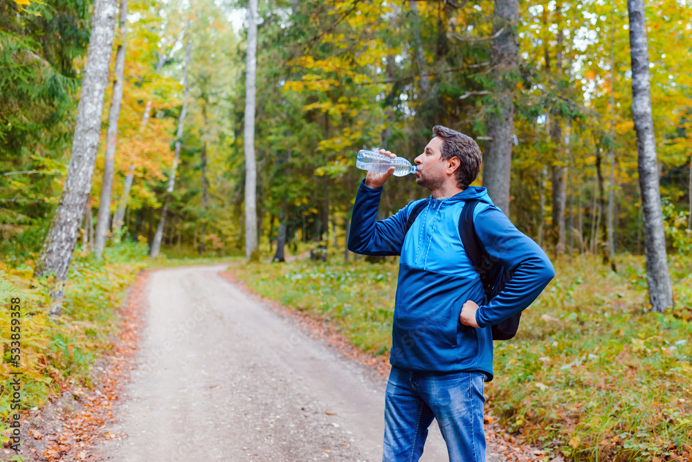 Fototapeta premium Middle age man hiker with backpack drinking water from the bottle, having break in autumn forest.