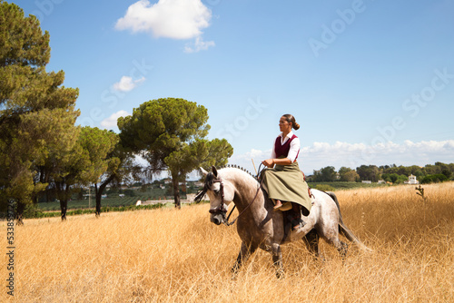 Young and beautiful Spanish woman on a Thoroughbred horse riding in the countryside in Spain. The woman is wearing a horse riding uniform. Thoroughbred and equine concept.