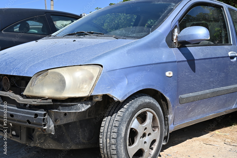 View on car damages in front part of the vehicle. Fender, halogen light and door back mirror are broken, their position is shifted and the paint is scratched. Forward plastic bumper is missing.