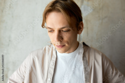 Portrait of handsome young serious man with blond hair dressed in casual beige shirt standing on gray textured wall looking down deep in thoughts, contemplating upon something in mind